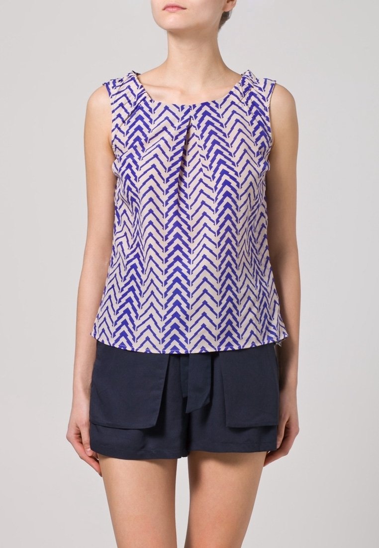 Bez rękawów bluzka w kremowym materiale z niebieskim wzorem w zygzak, z plisowanym dekoltem i luźnym krojem, noszona z ciemnymi szortami.