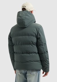 Gevoerde groene jas met een capuchon, voorzien van een geborduurd ontwerp en een gestructureerde stof. Draagt over lichtblauwe jeans.