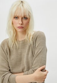 Beige gestrickter Pullover mit gerippter Textur und weiten Ärmeln. Der Stoff wirkt weich und lässig, ideal für entspannte Outfits.