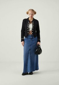 Jaqueta militar preta cropped com botões metálicos, blusa branca com pregas e saia longa de denim azul. Bolsa preta, sapatos planos.