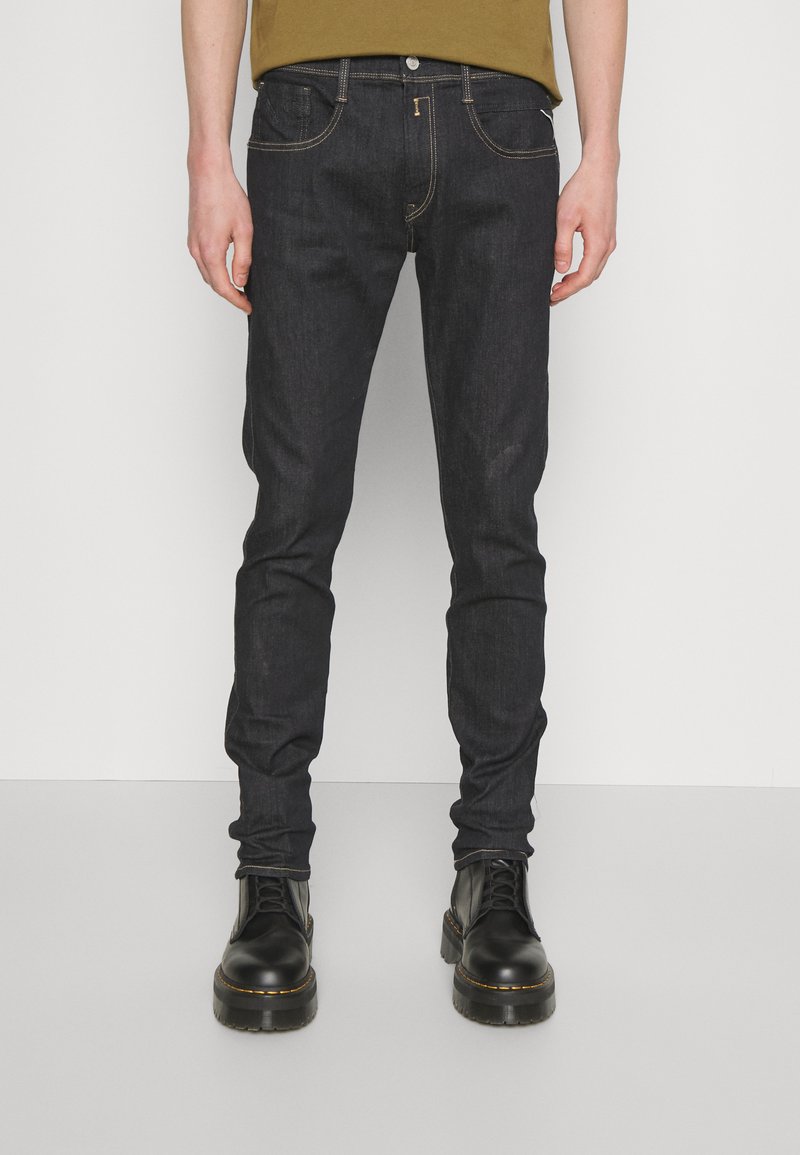 Jeans de mezclilla oscura con un ajuste Slim, que cuentan con costuras doradas, un diseño clásico de cinco bolsillos y un cierre de botón, combinados con botas negras hasta el tobillo.