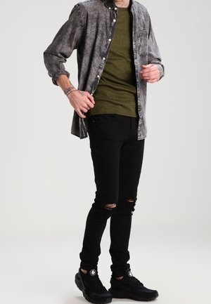 Graues Denim-Hemd über einem olivgrünen Strickpullover, kombiniert mit schwarzen, abgetragenen Jeans und schwarzen Sneakern. Minimalistisches Design.
