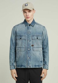 Ljusblå denimjacka med två bröstfickor, knäppning med knappar och krage. Har en logotyp patch på vänster bröst.