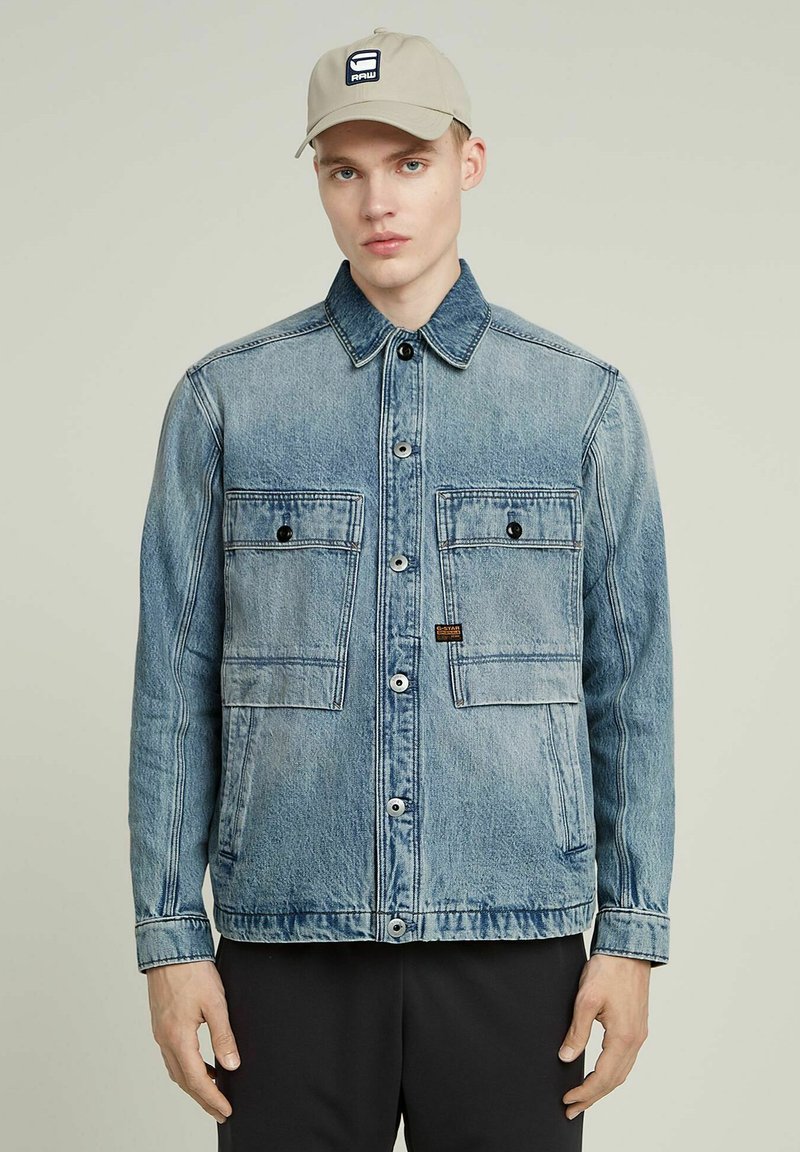 Ljusblå denimjacka med två bröstfickor, knäppning med knappar och krage. Har en logotyp patch på vänster bröst.