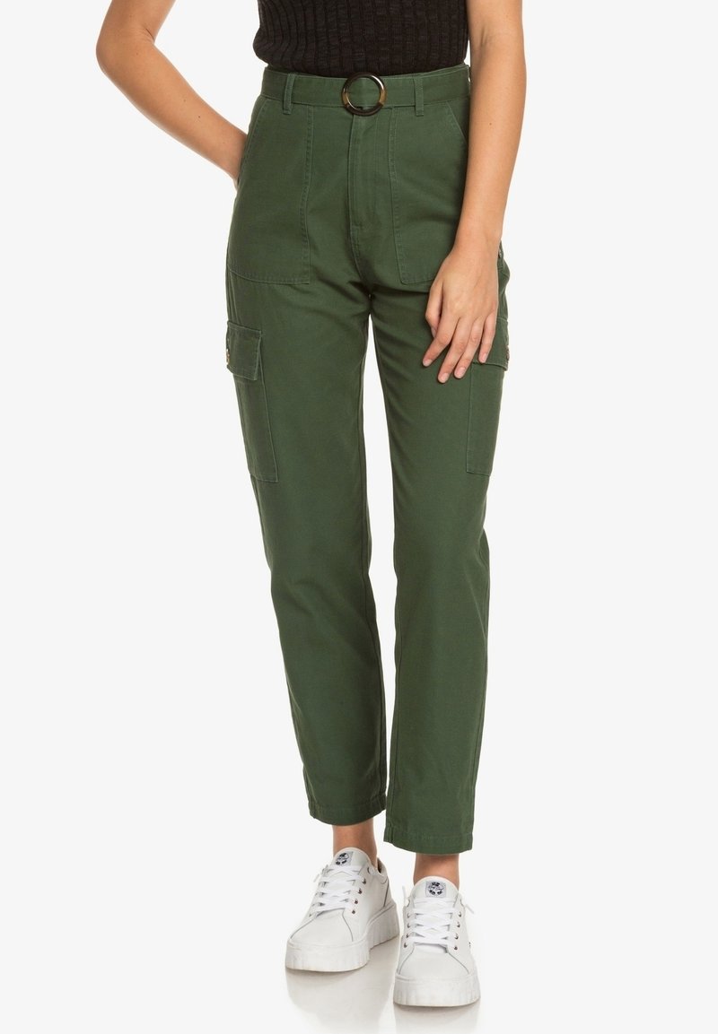Femme portant un pantalon cargo vert olive avec une boucle de ceinture ronde et des baskets blanches sur un fond uni.