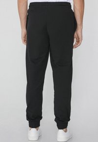 Zwarte joggerbroeken van zacht materiaal, met een elastische tailleband, gebundelde enkels en zijzakken. Schoeisel is witte sneakers.