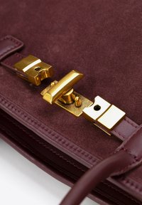 Burgunderfarbenes Wildleder-Handtasche mit goldfarbenen Beschlägen, einem strukturierten Verschlussmechanismus und detaillierter Naht an den Kanten.