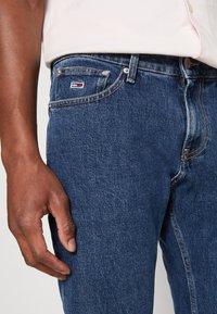 Modré džínové kalhoty s klasickým designem, s malým červeno-bílým logem na přední kapse a mírně opotřebovanou texturou.