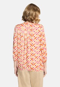 Femme aux cheveux blonds courts portant un chemisier à manches longues avec un motif géométrique rose, orange et rouge, associé à un pantalon beige, vue de dos.