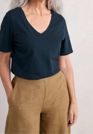 Person med gråt hår iført en mørk marineblå V-hals kortærmet skjorte, der er stoppet ned i højtaljede lysebrune linnedbukser med lommer.