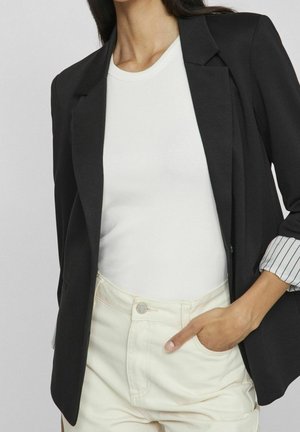 Femme portant un blazer noir aux manches rayées retroussées, une chemise blanche côtelée et un pantalon clair, une main dans la poche.