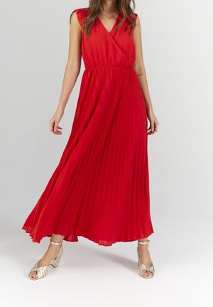 Kobieta w czerwonej, plisowanej sukni maxi bez rękawów, w złotych sandałach na obcasie, stojąca na jasnym, jednolitym tle.