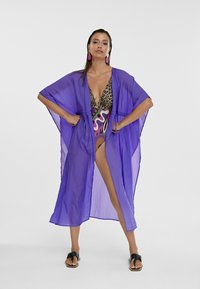 Copertura trasparente viola con maniche ampie, indossata sopra un costume da bagno con stampa leopardata. Include un profondo scollo a V e un design con apertura frontale.