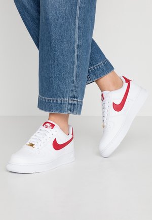 Personne portant des baskets blanches Nike Air Force 1 avec des accents rouges et un jean en denim bleu, debout sur un sol gris clair uni.