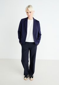 Blazer blu navy con rever affilati, indossato su una camicia bianca. Abbinato a pantaloni larghi scuri e sneakers nere e crema. Design minimalista.