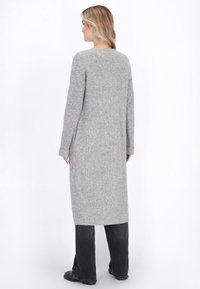 Donna che indossa un lungo cardigan grigio a coste con maniche a risvolto, abbinato a pantaloni neri a gamba larga e scarpe nere, in piedi di spalle.
