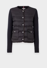 Zwarte pufferjas met een gebreide marineblauwe lijf en mouwen. Voorzien van drukknoopsluitingen, twee voorkruiszakken en geribde manchetten.