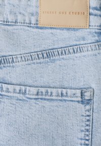 Tissu en denim bleu clair avec une poche arrière et une étiquette en cuir beige cousue au-dessus, embossée avec le texte "STREET ONE STUDIO."