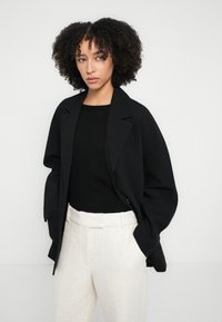 Blazer negru supradimensionat cu rever cu notch, asortat cu un pulover negru și pantaloni albi texturați, cu talie medie și picioare largi.