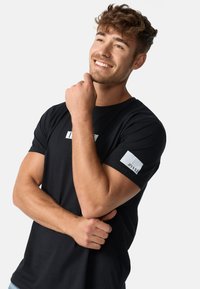 Čierne tričko s krátkym rukávom s bielym obdĺžnikovým logom na hrudi a bielou štítkou s nápisom „1993“ na ľavom rukáve. Mäkká látka.