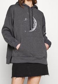 Sweat à capuche gris avec une poche avant, arborant un motif de lune et d'étoile. Fini avec une capuche à cordon de serrage et des poignets et ourlet côtelés.