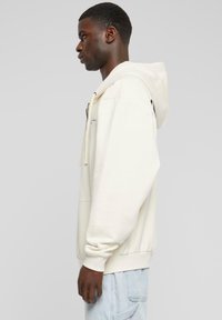 Cream hoodie with a drawstring and front pocket; ribbed cuffs and hem; soft texture; worn over light blue jeans. Side profile view.