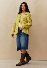 Žlutý pletený cardigan s texturovanými kabelkami, oversized střih, zapínání na knoflíky. Nosený přes bílý top a spárovaný s modrými džínovými šortkami a hnědými botami.