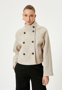 Beige taillierte Doppelreiher-Jacke mit hohem Kragen, schwarzen Knöpfen, seitlichen Taschen und glattem, wollähnlichem Material.