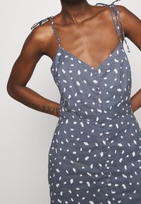 Niebieska sukienka z białymi wzorami w płatki, cienkie ramiączka związane na ramionach, dekolt w kształcie litery V, zapięcie na guziki z przodu oraz dopasowany krój.
