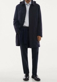 Abrigo azul marino con capucha, mangas largas y bolsillos delanteros; se lleva sobre una camisa azul claro y pantalones oscuros; combinado con zapatos negros.