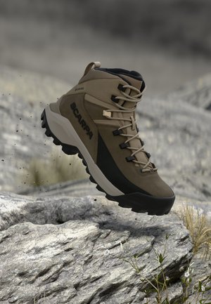 Bruine en zwarte Scarpa-wandelschoen zweeft boven rotsachtig terrein met kleine planten en een vage achtergrond.