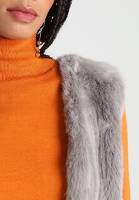 Maglione arancione a collo alto in tessuto a coste, abbinato a un morbido gilet in pelliccia sintetica grigia con superficie strutturata e senza chiusure visibili.