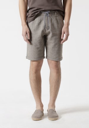 Hombre con pantalones cortos beige con dobladillo enrollado con cordón blanco y azul, camisa marrón oscuro y alpargatas beige sin cordones, de pie sobre fondo blanco.