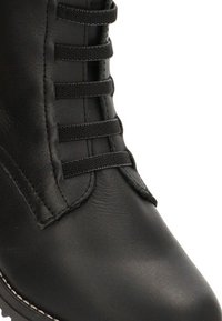 Botín de cuero negro que presenta una punta redondeada, cordones decorativos y costuras blancas en la suela para un detalle adicional.