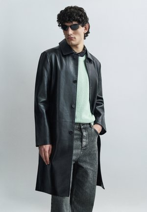 Svart läderkappa med slagkrage, knäppning fram och sidofickor, bärs över en mintgrön tröja och grå denimbyxor. Svarta solglasögon.