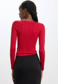 Maglietta rossa a maniche lunghe con un design aderente e una texture scintillante, caratterizzata da un ampio scollo e orlo rifinito, abbinata a una gonna nera.