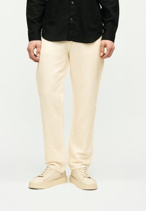 Homme portant un pantalon crème, des baskets crème assorties et une chemise noire à boutons, debout devant un fond blanc.