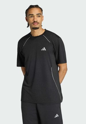 Mand med flettet hår iført en sort Adidas trøje og shorts med reflekslogo og hvide detaljer, stående foran en ensfarvet baggrund.