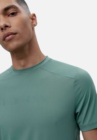 Gaiši zaļš sporta krekls ar īsām piedurknēm un apaļu apkakli, izgatavots no gludas, stiepjamas auduma un ar diskrētu zīmola atzīmi priekšpusē.