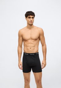 Homem sem camisa com cabelo curto e escuro, vestindo boxers pretos da Calvin Klein, em pé contra um fundo cinza claro liso.