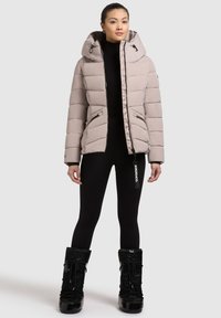 Beige színű puffermellény kapucnival és cipzárral, steppelt dizájnnal és oldalzsebekkel. Fekete leggings-szel és fekete csizmával viselve.