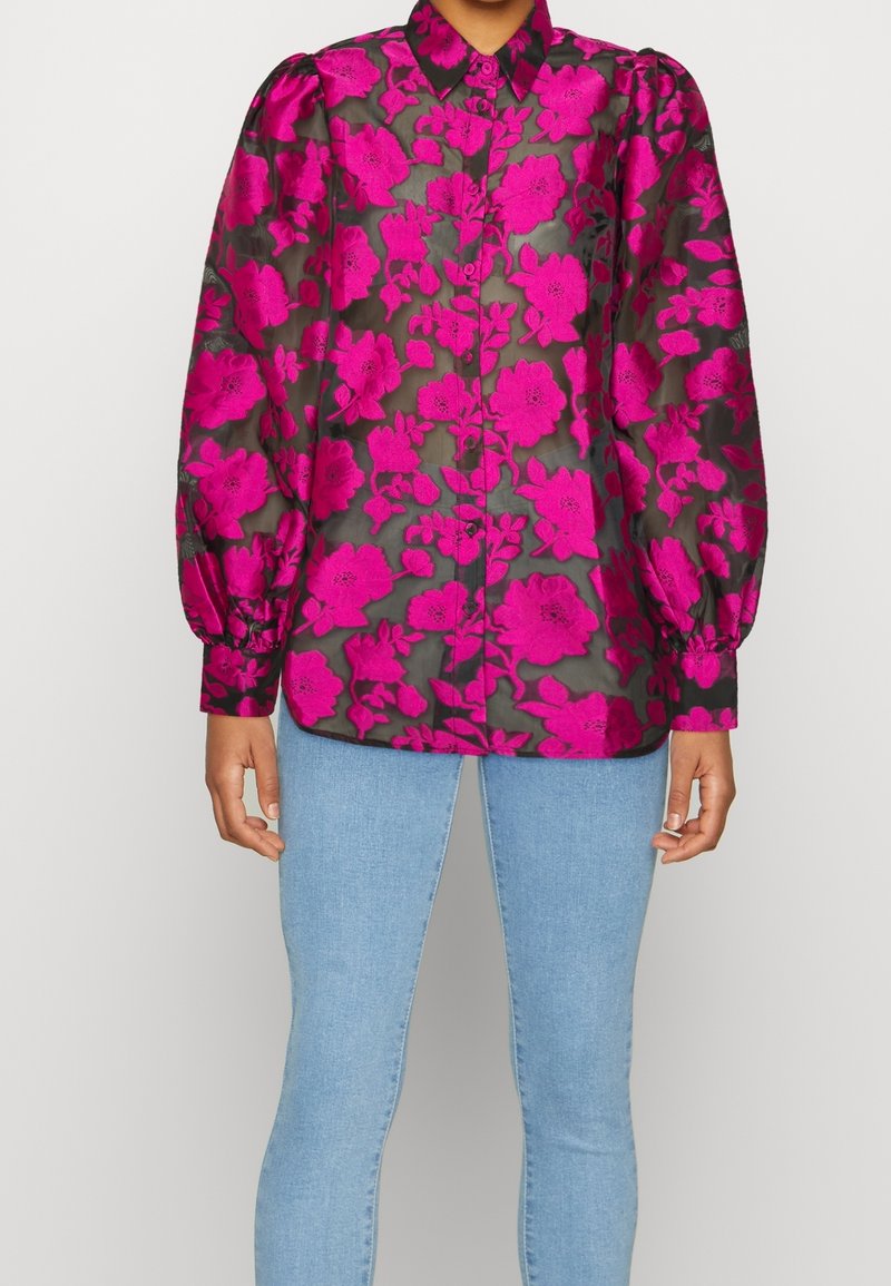 Blouse noire transparente avec grand motif floral rose vif, manches longues bouffantes, poignets boutonnés et jean bleu clair ajusté.