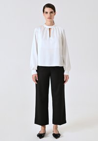 Weißes Langarmblusenshirt mit Schlüsselloch-Ausschnitt und gerafftem Halsauschnitt, kombiniert mit schwarzen weiten Hosen und schwarzen spitzen Loafers.