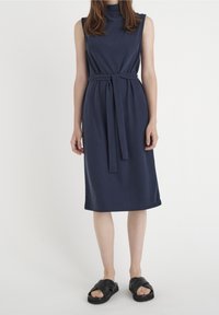 Granatowa sukienka bez rękawów z wysokim dekoltem, zaostrzona w talii za pomocą materiałowego paska, o długości midi. Połączona z czarnymi sandałami na paski.