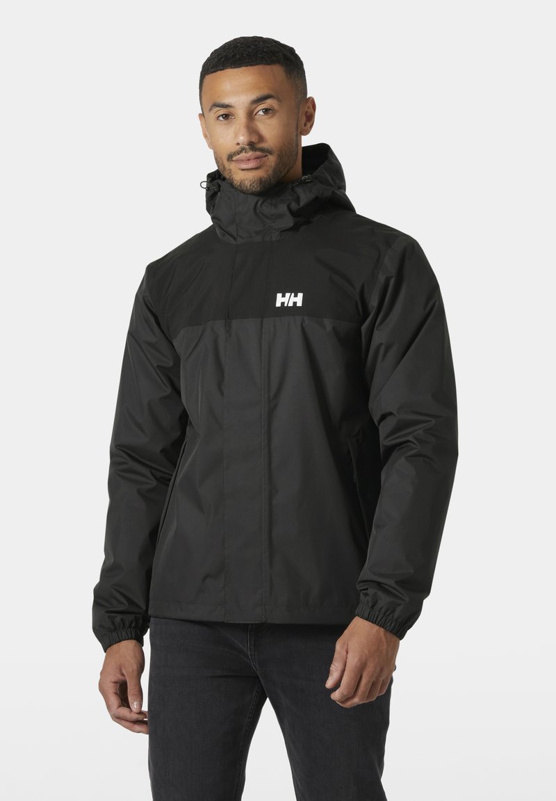Homme portant une veste à capuche noire Helly Hansen avec une fermeture éclair devant et des poignets élastiques, debout devant un fond gris clair uni.