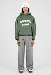 Hombre con un gorro de lana blanco, sudadera verde oliva con el texto "PROHIBIDO BERLÍN", pantalones anchos grises y zapatos bicolores, de pie contra un fondo blanco.