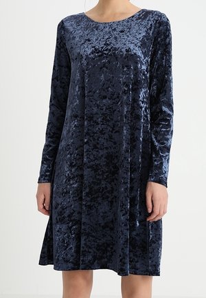 Femme portant une robe en velours froissé bleu foncé, longueur genoux, manches longues et col rond.