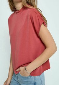 Haut rouge à manches courtes avec col montant, en tissu doux. Présente une coupe décontractée et est rentré dans un denim bleu clair.