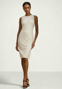 Vestido bege sem mangas com uma silhueta ajustada, comprimento até ao joelho, textura suave e design minimalista. Combinado com saltos altos pretos com tiras.