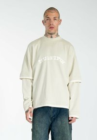 Off-White übergroßer Sweatshirt mit strukturierter Oberfläche, mit dem Schriftzug "MULTIPLY" in kontrastierender Schrift, kombiniert mit blauen Jeans.